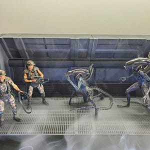 May include: A diorama featuring two articulated soldier action figures in camouflage gear and two black, elongated extraterrestrial creature figures. The scene is set against a dark, industrial-style wall panel with a grated floor and wispy smoke.