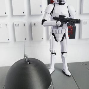 May include: A white Stormtrooper action figure holding a black blaster rifle, standing in front of a white wall with rectangular panels. A black, dome-shaped object with a silver antenna is in the foreground. The figure is wearing a white helmet and armor.