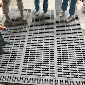 May include: Grey plastic modular flooring tiles with a slotted design. The tiles are arranged in a grid pattern, with visible measurement markings along the edges. Several pairs of legs and feet are visible, standing on the tiles.