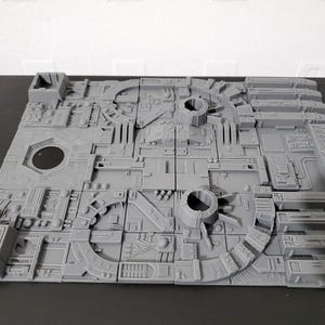 May include: A gray, modular, 3D-printed terrain piece with intricate details. The design features circular and rectangular elements, along with various mechanical components. The surface is textured, and the piece is likely for tabletop gaming or model building.
