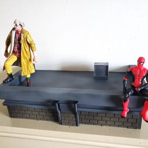 Peut inclure: Un socle de présentation diorama en forme de toit gris avec un mur avant en briques, deux tuyaux verticaux et une petite bouche d'aération carrée. Deux figurines sont placées sur la structure : une portant un manteau jaune et une en costume rouge et noir.