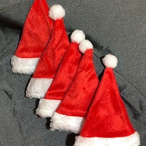 May include: Five red Santa hats with white faux fur trim and white pom-poms. The hats are arranged in a diagonal row against a gray background. Festive holiday decor.