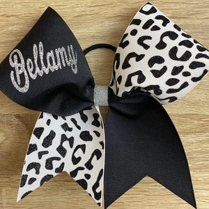 May include: A hair bow featuring a black ribbon side with the name Bellamy in silver glitter and a white ribbon side with a black leopard print pattern. The centre is cinched with a silver glitter band and includes a black elastic hair tie.