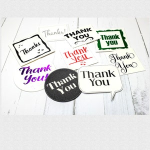 May include: Assortment of thank you cards with different designs and fonts. The cards are white, black, green, and purple with black, red, and white lettering. The cards are on a white wooden surface.