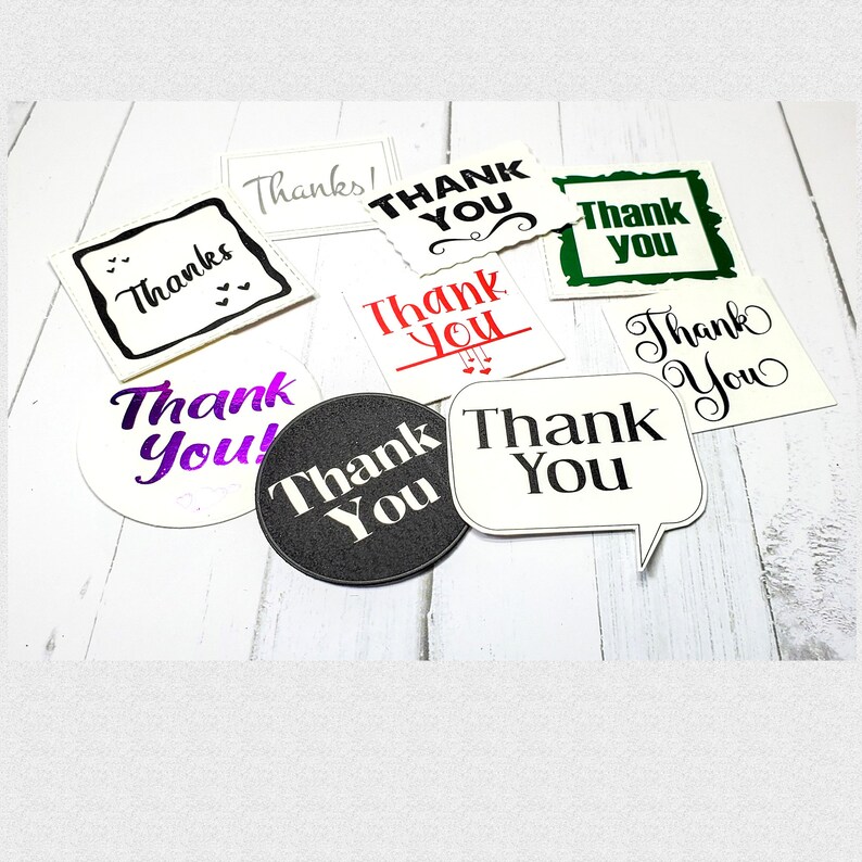May include: Assortment of thank you cards with different designs and fonts. The cards are white, black, green, and purple with black, red, and white lettering. The cards are on a white wooden surface.