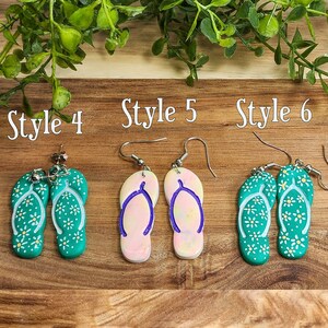 May include: Four pairs of dangle earrings with a flip-flop design. The earrings are made of polymer clay and feature different colors and patterns. The first pair is green with white flowers, the second pair is pink and white with a purple outline, the third pair is green with white flowers, and the fourth pair is green with white flowers.