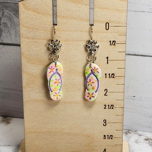 May include: A pair of dangle earrings featuring flip-flop charms with a floral design in shades of pink, yellow, and green. The earrings are displayed on a wooden stand with a ruler measuring up to 4 inches.