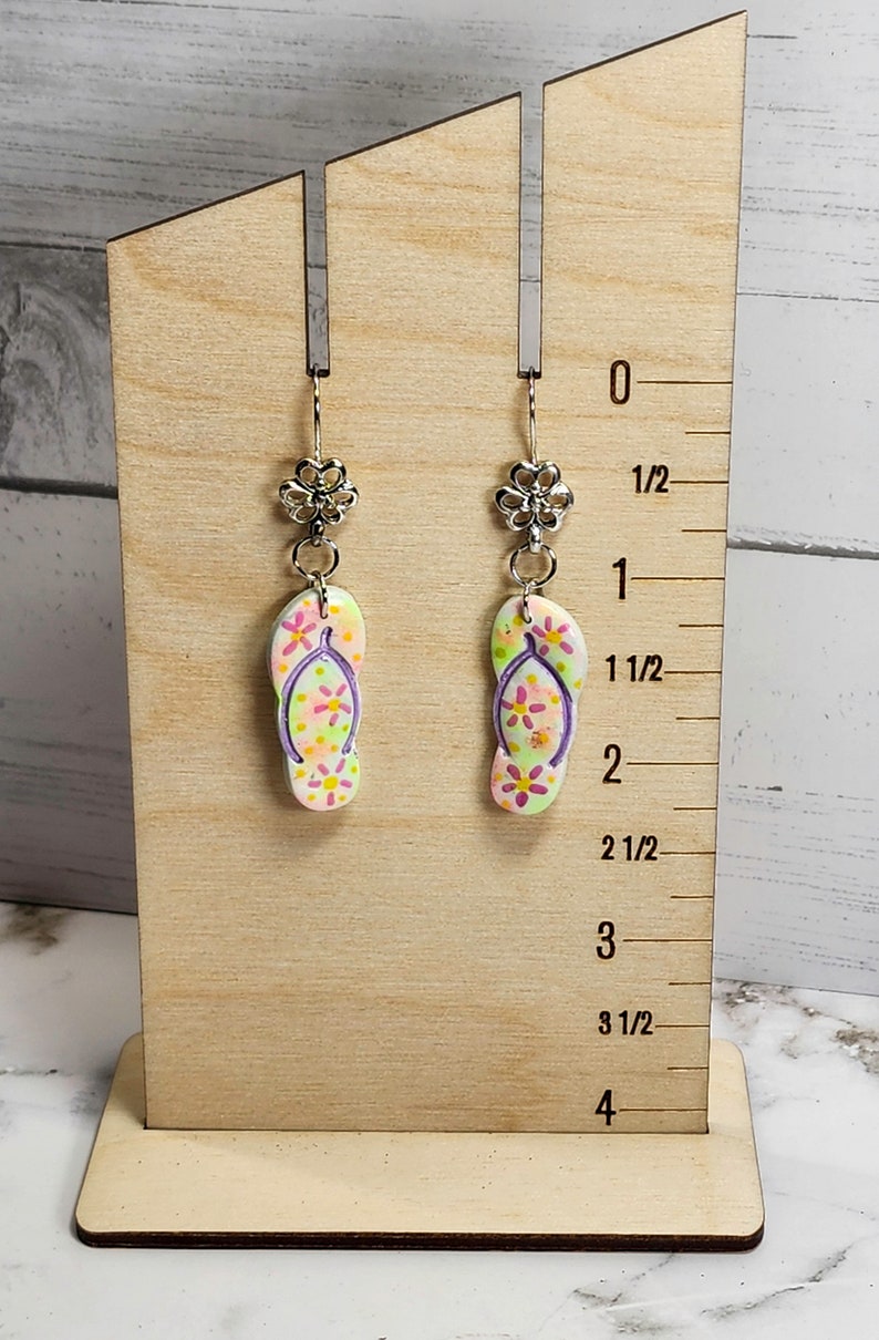 May include: A pair of dangle earrings featuring flip-flop charms with a floral design in shades of pink, yellow, and green. The earrings are displayed on a wooden stand with a ruler measuring up to 4 inches.