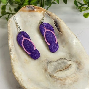May include: Purple glitter flip flop earrings with pink straps. The earrings are hanging from silver hooks.
