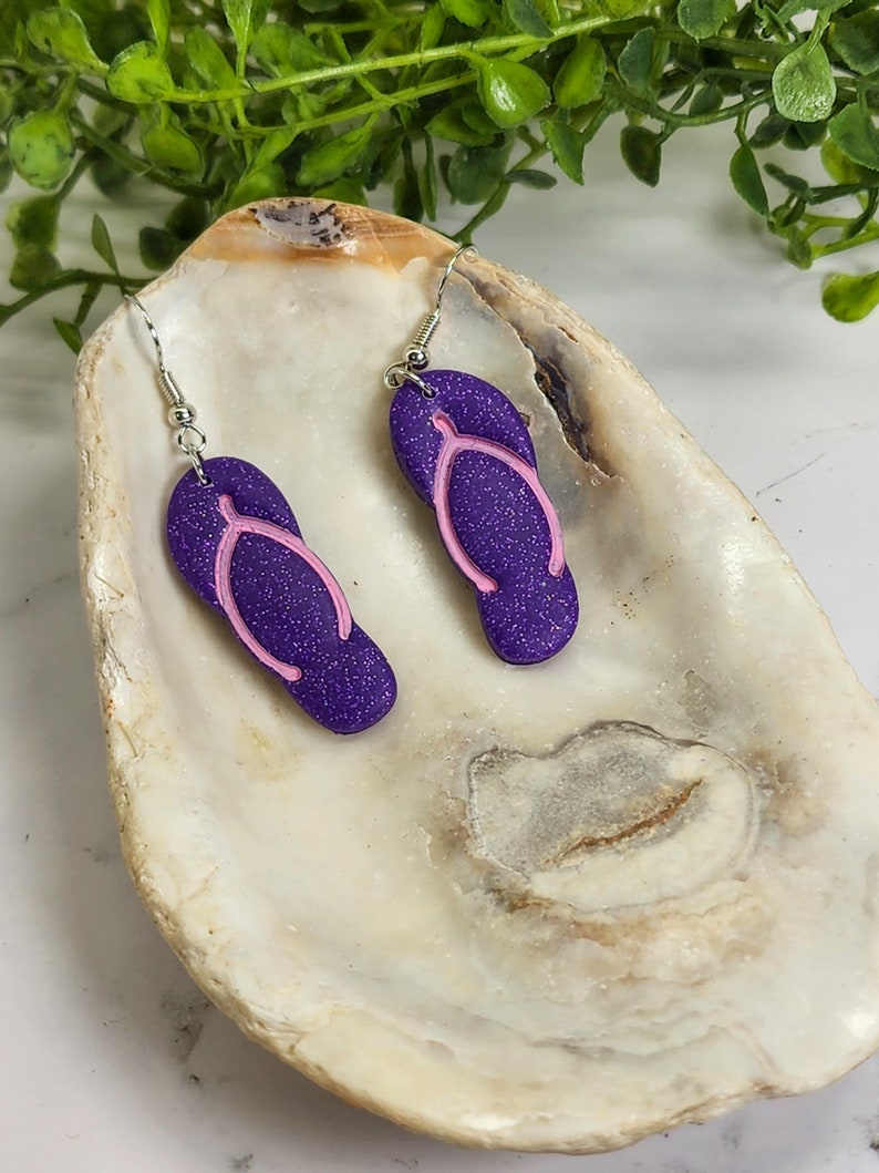 May include: Purple glitter flip flop earrings with pink straps. The earrings are hanging from silver hooks.