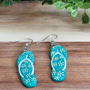 May include: A pair of teal blue flip-flop earrings with white daisy accents. The earrings have silver hooks.