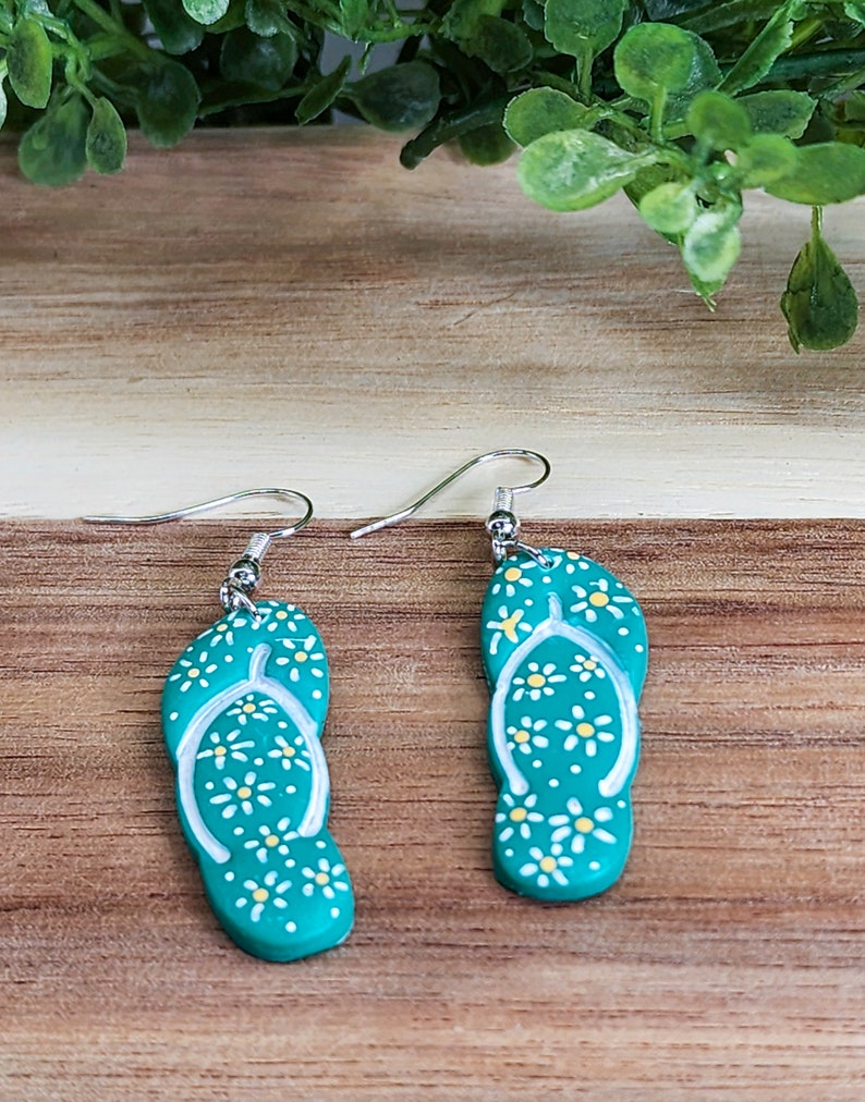 May include: A pair of teal blue flip-flop earrings with white daisy accents. The earrings have silver hooks.