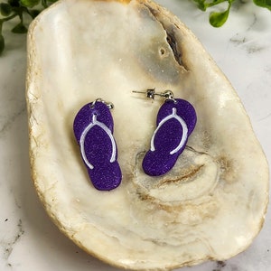 May include: A pair of purple flip-flop earrings with white outlines. The earrings are displayed on a white seashell.