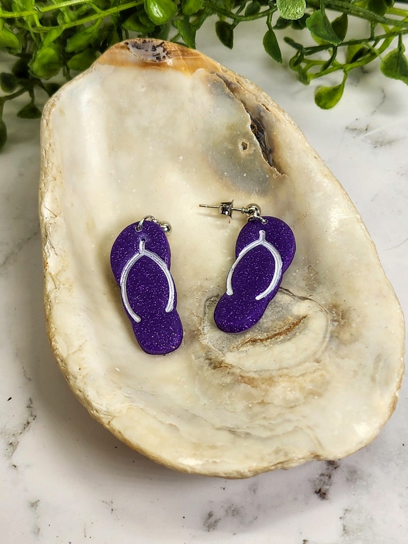 May include: A pair of purple flip-flop earrings with white outlines. The earrings are displayed on a white seashell.