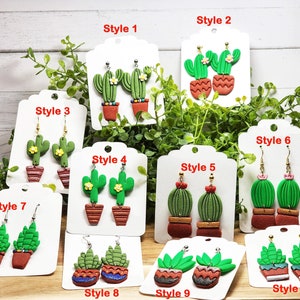 May include: Ten pairs of cactus-themed dangle earrings displayed on white cards, labeled Style 1 through Style 10. Each pair features green textured cacti in terracotta-colored pots, some with small flower accents, hanging from gold or silver hooks.