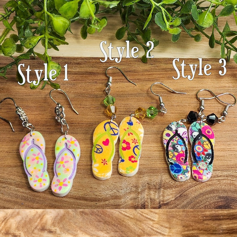 May include: Three pairs of flip-flop earrings in different styles. The first pair is yellow with peace signs and flowers, the second pair is yellow with flowers, and the third pair is black and white with a yin-yang symbol and flowers.