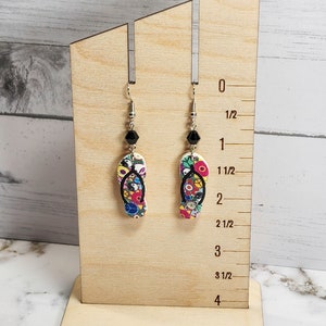 May include: A pair of colorful flip-flop earrings with black beads hanging from silver hooks. The earrings are displayed on a wooden stand with a ruler measuring up to 4 inches.