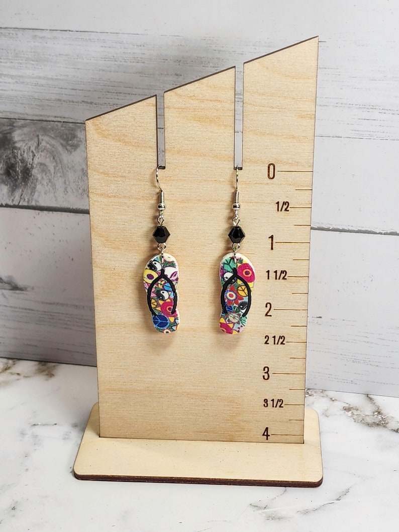 May include: A pair of colorful flip-flop earrings with black beads hanging from silver hooks. The earrings are displayed on a wooden stand with a ruler measuring up to 4 inches.