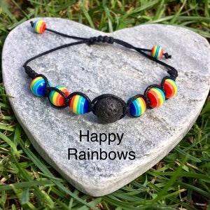 May include: A black string bracelet with rainbow coloured beads and a black lava rock bead. The bracelet is on a heart-shaped stone with the text "Happy Rainbows" written on it.