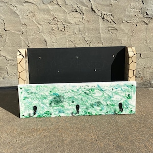 Puede incluir: Un porta llaves de madera con tres ganchos. El panel frontal está pintado con pintura marmoleada verde y blanca. El panel trasero es negro y tiene un patrón geométrico.