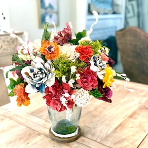 May include: A colorful floral arrangement in a clear glass vase with a wooden base. The bouquet features a variety of paper flowers in shades of red, orange, yellow, white, and blue. The vase sits on a light-colored wooden table.