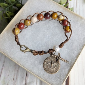 May include: A brown leather bracelet with multi-colored beads and a metal charm with a dove and cross. The bracelet is in a white box.
