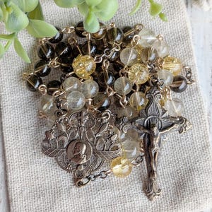 May include: A rosary with brown and yellow beads, a decorative metal centerpiece, and a crucifix. The beads are a mix of clear and opaque, with a metallic chain. The rosary is displayed on a beige cloth.