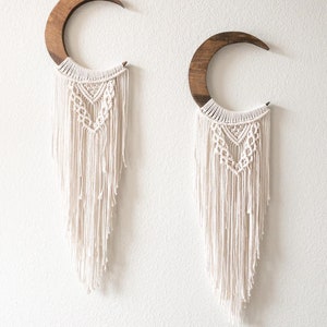 May include: Two crescent moon wall hangings. Each features a dark wood crescent shape with a cream-colored macrame design. The macrame has a geometric pattern at the top and long fringe. The wall hangings are mounted on a white wall.