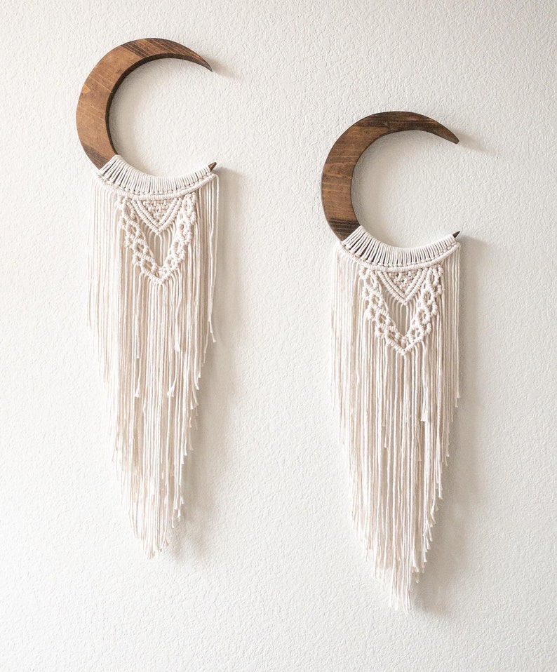 May include: Two crescent moon wall hangings. Each features a dark wood crescent shape with a cream-colored macrame design. The macrame has a geometric pattern at the top and long fringe. The wall hangings are mounted on a white wall.