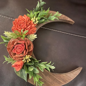 May include: A wooden crescent moon wall hanging adorned with orange and brown flowers and green foliage. The flowers are clustered on the left side of the moon, with the leaves and small yellow flowers filling the gaps.