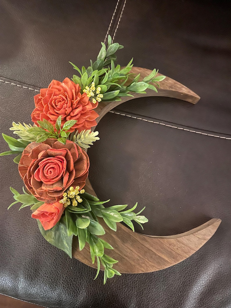 May include: A wooden crescent moon wall hanging adorned with orange and brown flowers and green foliage. The flowers are clustered on the left side of the moon, with the leaves and small yellow flowers filling the gaps.
