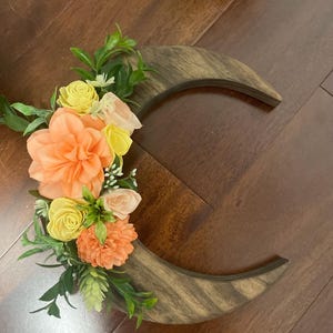 May include: A wooden crescent moon decoration with a floral arrangement. The flowers are in shades of peach, yellow, and pink, with green leaves. The wood has a natural grain pattern.