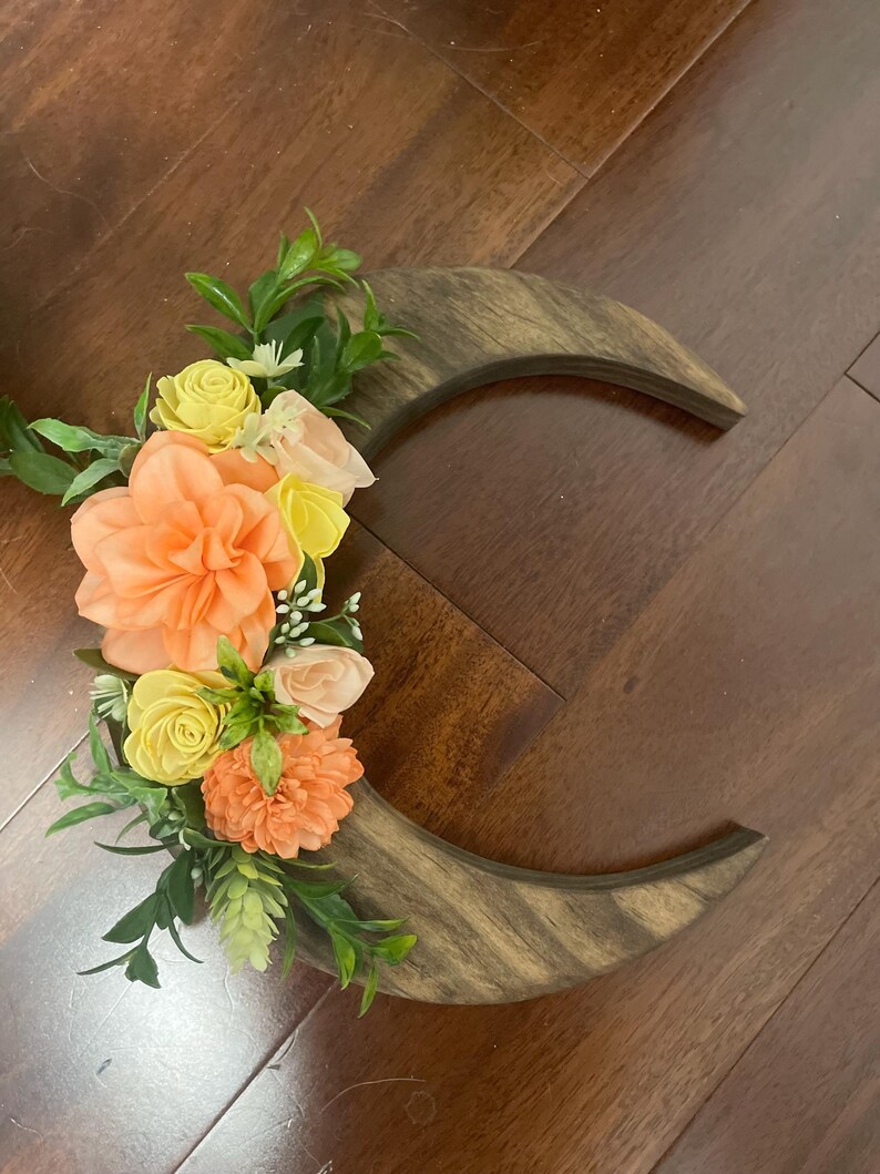 May include: A wooden crescent moon decoration with a floral arrangement. The flowers are in shades of peach, yellow, and pink, with green leaves. The wood has a natural grain pattern.