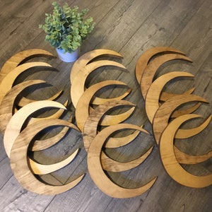 May include: A collection of wooden crescent moon shapes in varying shades of brown. The moons are arranged on a wooden floor, with a small white pot of greenery in the background. The moons appear to be cut from wood and have a natural finish.