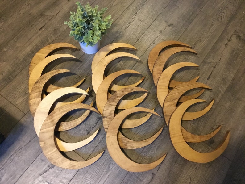 May include: A collection of wooden crescent moon shapes in varying shades of brown. The moons are arranged on a wooden floor, with a small white pot of greenery in the background. The moons appear to be cut from wood and have a natural finish.