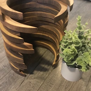 May include: A unique wooden side table with a spiral design. The table is constructed from curved, light brown wooden pieces stacked to create a cylindrical shape. A small potted plant with green leaves sits beside the table.
