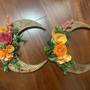 May include: Two wooden crescent moon wall hangings. Each moon is decorated with colorful faux flowers in shades of orange, yellow, and burgundy, with green foliage and berries. The moons are made of stained wood.