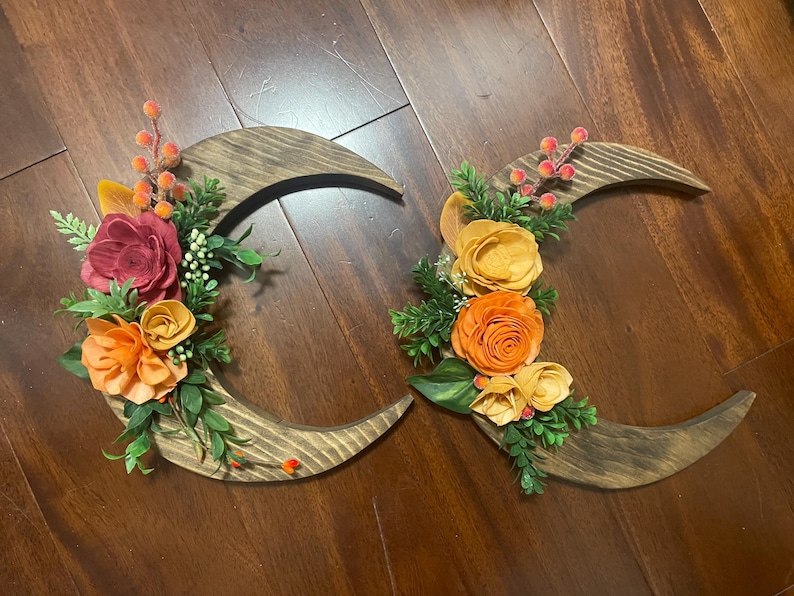 May include: Two wooden crescent moon wall hangings. Each moon is decorated with colorful faux flowers in shades of orange, yellow, and burgundy, with green foliage and berries. The moons are made of stained wood.