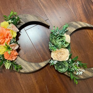 May include: Wooden crescent moon wall decor with floral accents. The moons are made of dark wood and adorned with colorful flowers in shades of orange, yellow, and green, creating a rustic and natural aesthetic.
