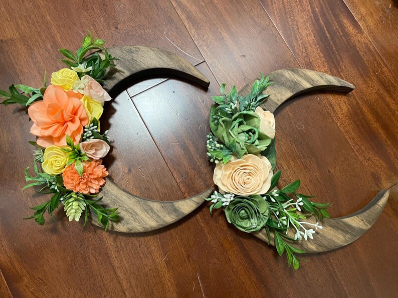 May include: Wooden crescent moon wall decor with floral accents. The moons are made of dark wood and adorned with colorful flowers in shades of orange, yellow, and green, creating a rustic and natural aesthetic.