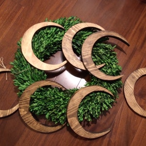 May include: Wooden crescent moon shapes in various sizes and shades of brown are arranged on a green wreath. The moons have a natural wood grain finish. The wreath is made of small, dense green leaves. The arrangement is set on a wooden floor.