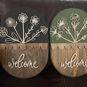 May include: Two oval wooden welcome signs. One is dark green with light wood flowers, the other is black with light wood flowers. Both have a scalloped edge and the word "welcome" in cursive.