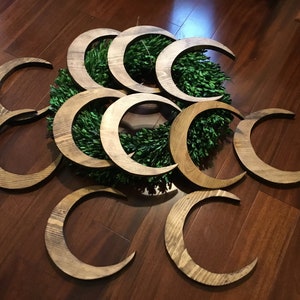 May include: Wooden crescent moon decorations in various sizes, arranged around a green wreath. The moons have a natural wood grain finish and are displayed on a dark wood surface. The overall theme is celestial and rustic.