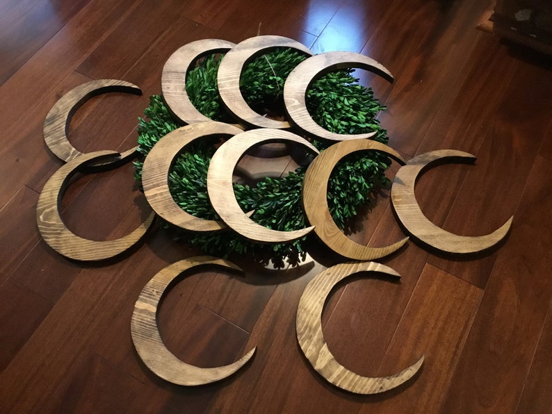 May include: Wooden crescent moon decorations in various sizes, arranged around a green wreath. The moons have a natural wood grain finish and are displayed on a dark wood surface. The overall theme is celestial and rustic.