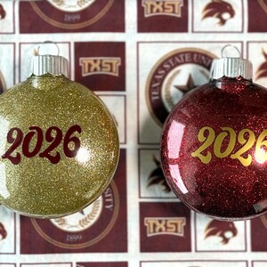 May include: Two round glass ornaments with silver caps. The left ornament is covered in gold glitter with the year 2026 printed in dark red. The right ornament is covered in deep red glitter with the year 2026 printed in gold lettering.