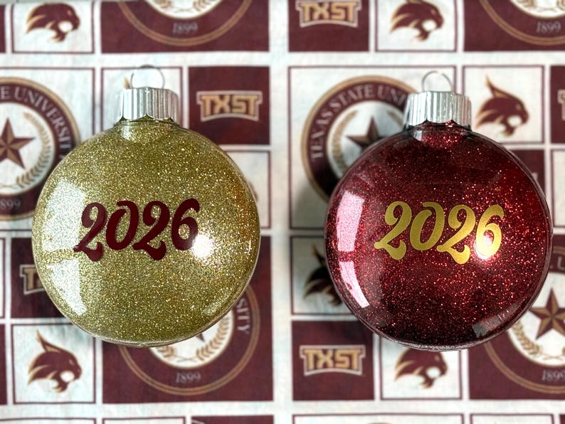 May include: Two round glass ornaments with silver caps. The left ornament is covered in gold glitter with the year 2026 printed in dark red. The right ornament is covered in deep red glitter with the year 2026 printed in gold lettering.
