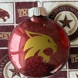 May include: A round, red glitter-filled Christmas ornament featuring a gold-colored bobcat head logo on the front. The ornament has a silver-toned metal cap at the top with a small wire loop for hanging, creating a festive holiday decoration.