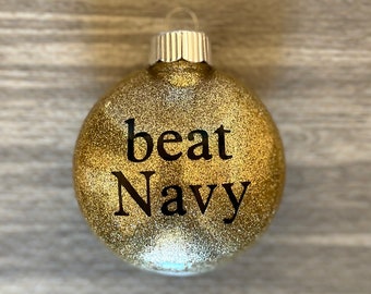 May include: Gold glitter Christmas ornament with the text "beat Navy" in black lettering.