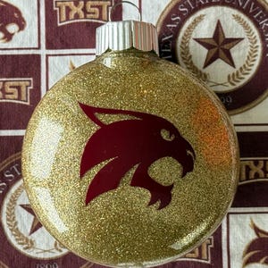 May include: A round, clear glass Christmas ornament filled with gold glitter. The front features a maroon bobcat head logo. A silver metal cap with a small wire loop sits at the top of the ornament, allowing it to be hung on a tree or display.