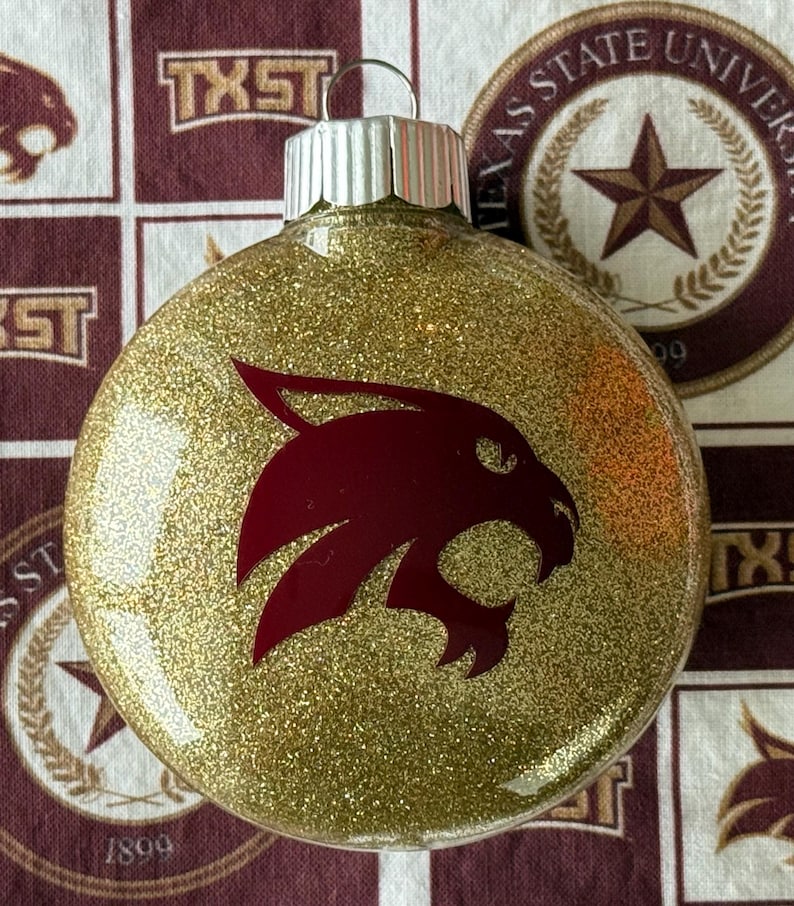 May include: A round, clear glass Christmas ornament filled with gold glitter. The front features a maroon bobcat head logo. A silver metal cap with a small wire loop sits at the top of the ornament, allowing it to be hung on a tree or display.